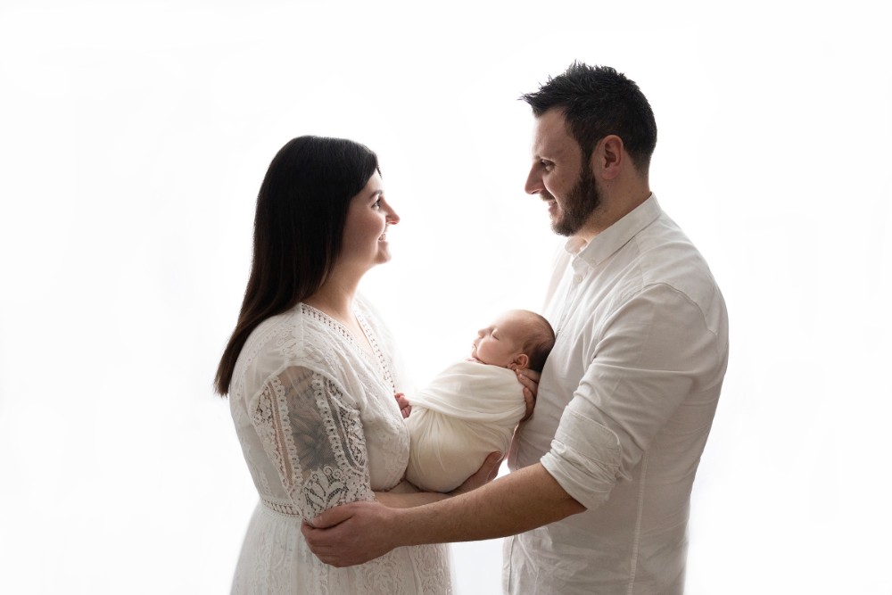 Parents lovingly holding swaddled baby girl in white, smiling family portrait, professional Essex photography