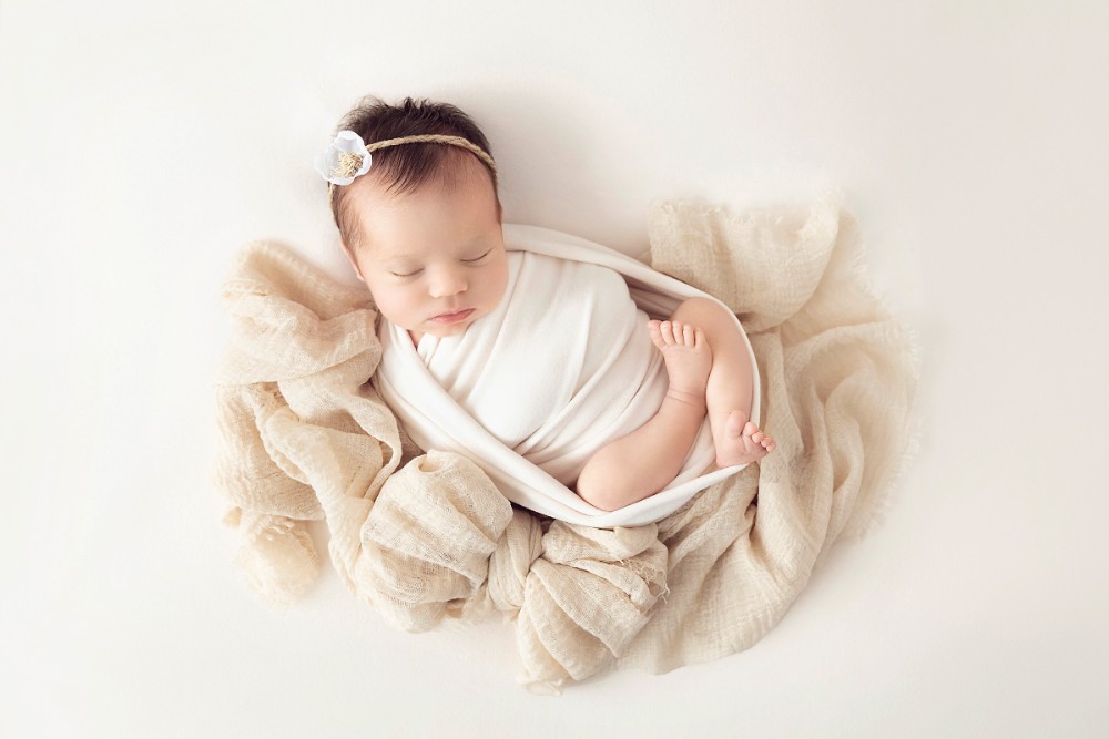 Newborn girl wrapped in cream blanket with floral headband, peacefully sleeping in Essex