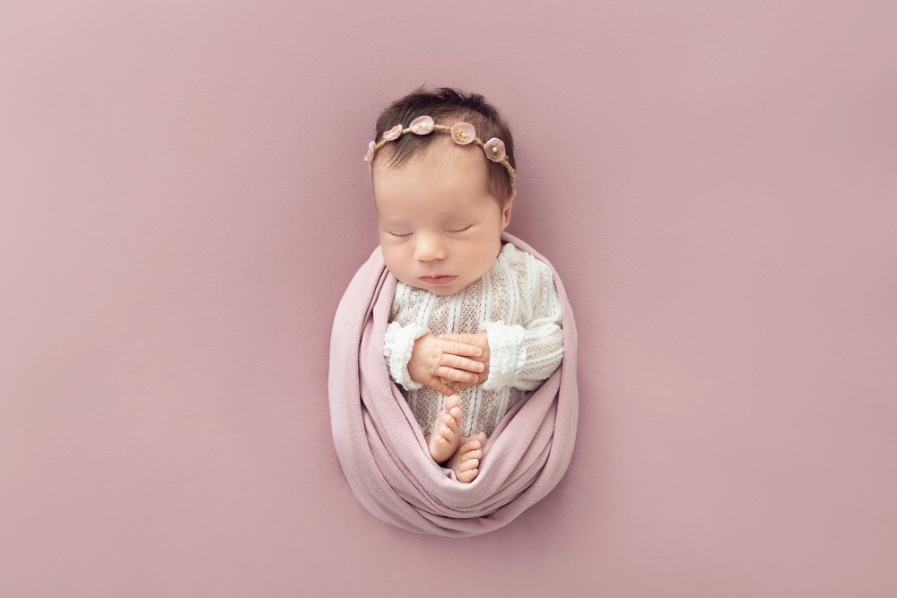 Newborn in pink lace romper and floral headband, posed on pastel background in Essex