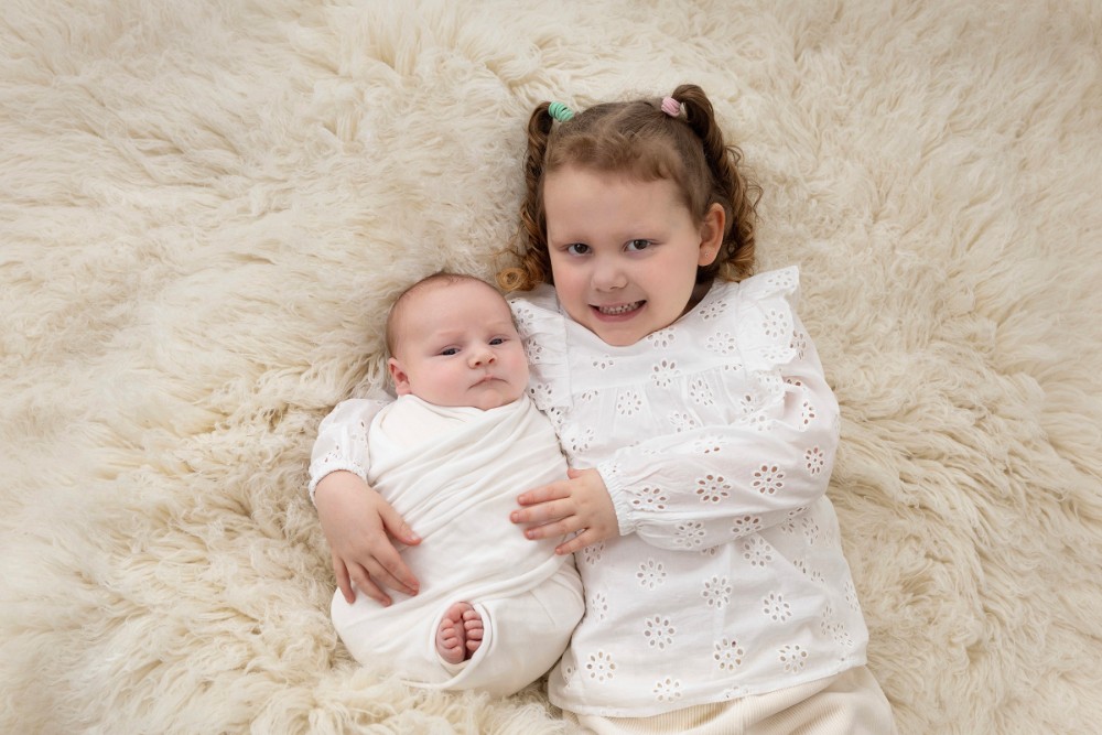 Big sister cuddles swaddled newborn brother on white fur, sweet sibling bond portrait in Essex