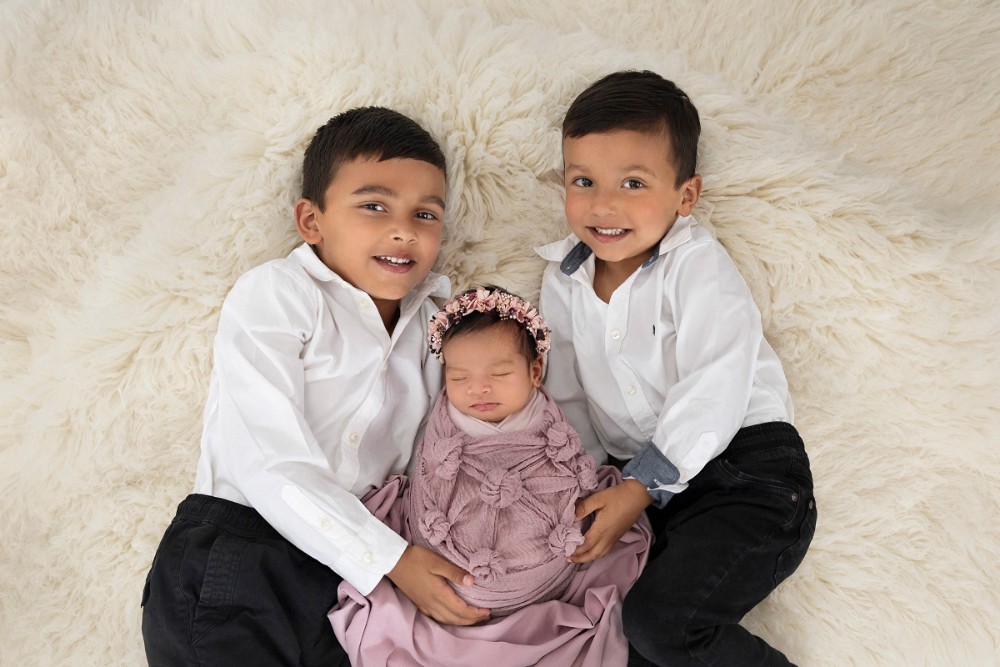 Two brothers holding newborn sister in pink wrap, sweet siblings portrait on soft fur in Essex