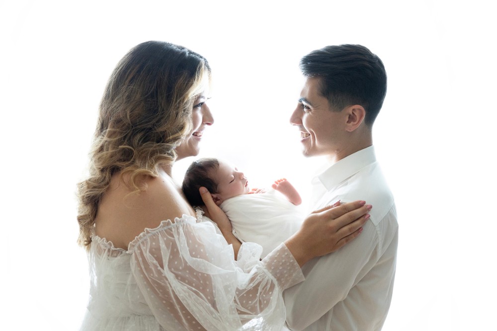 Mom and dad gazing at swaddled newborn in white Essex studio, heartwarming family portrait