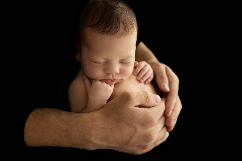 Father cradling sleeping newborn against black backdrop, capturing a timeless Essex baby portrait