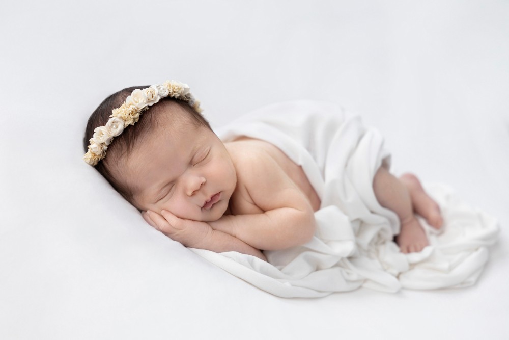 Newborn girl sleeping on white drape with floral headband, serene Essex professional photoshoot