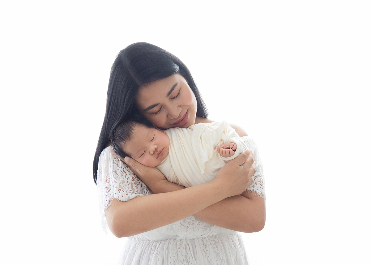Mother gently holding sleeping newborn baby in white dress during calm natural newborn photography session in Colchester Essex.