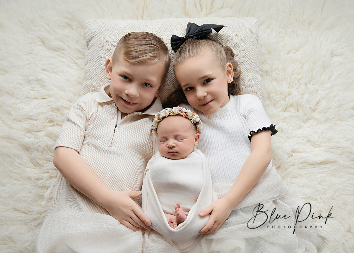 Siblings holding newborn baby gently wrapped in white wrap during in-home photoshoot in Essex