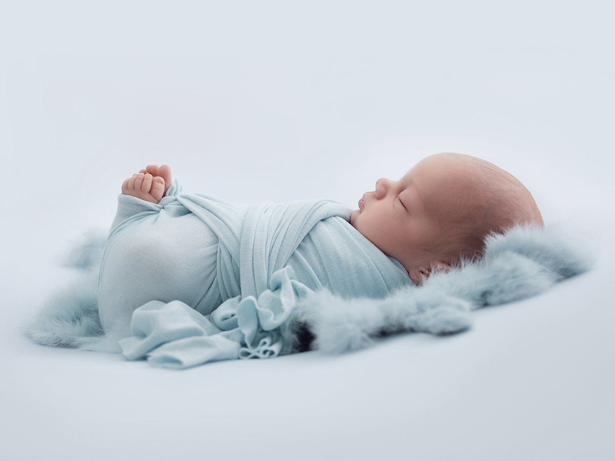 Sleeping newborn baby wrapped in soft blue fabric resting on textured blanket during professional newborn photography session in Halstead Essex.