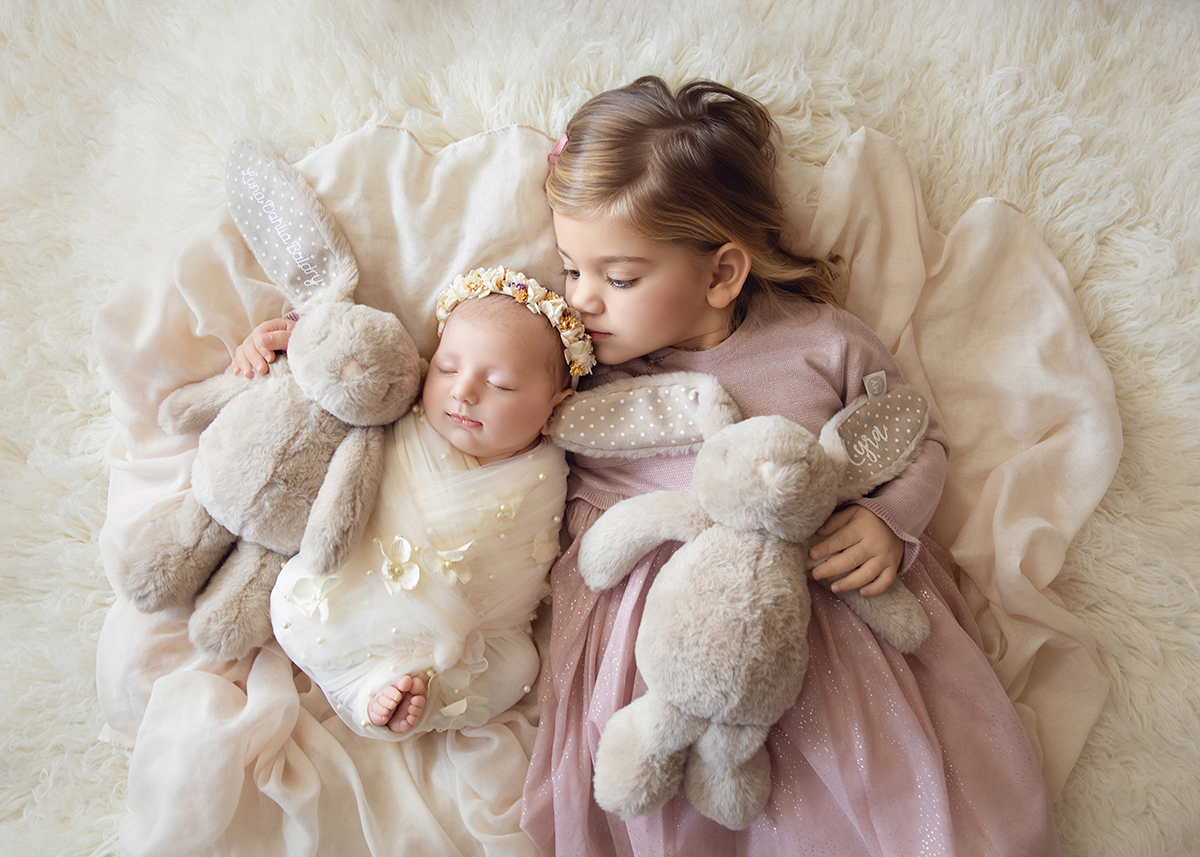 Older sister wearing a soft pastel dress cuddling her newborn sibling during an in-home family photography session in Essex.
